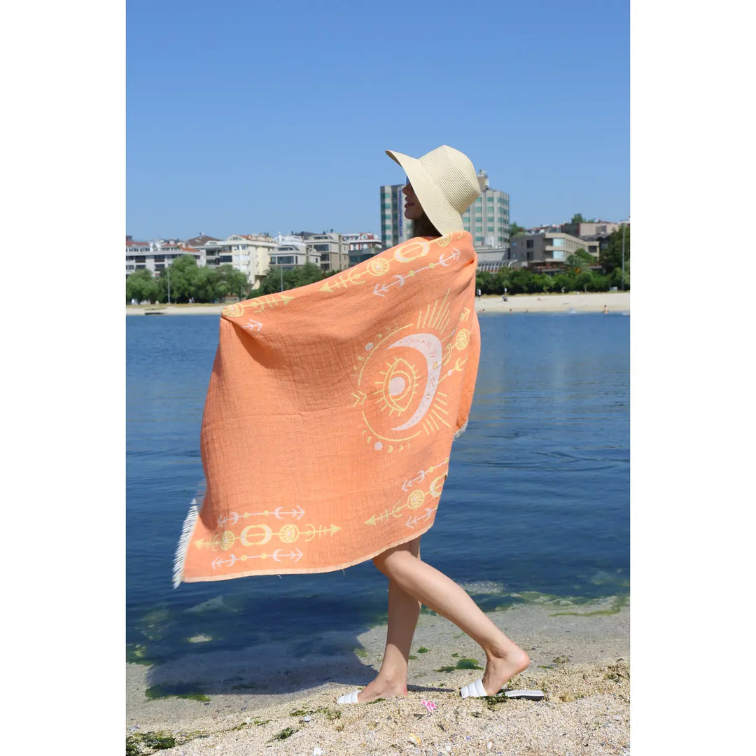 A person in a sunhat stands on a sandy beach by the water, wrapped in the Orange Seeing Eye Turkish Towel from turkanhome.com. Buildings and greenery are visible across the water in the background.