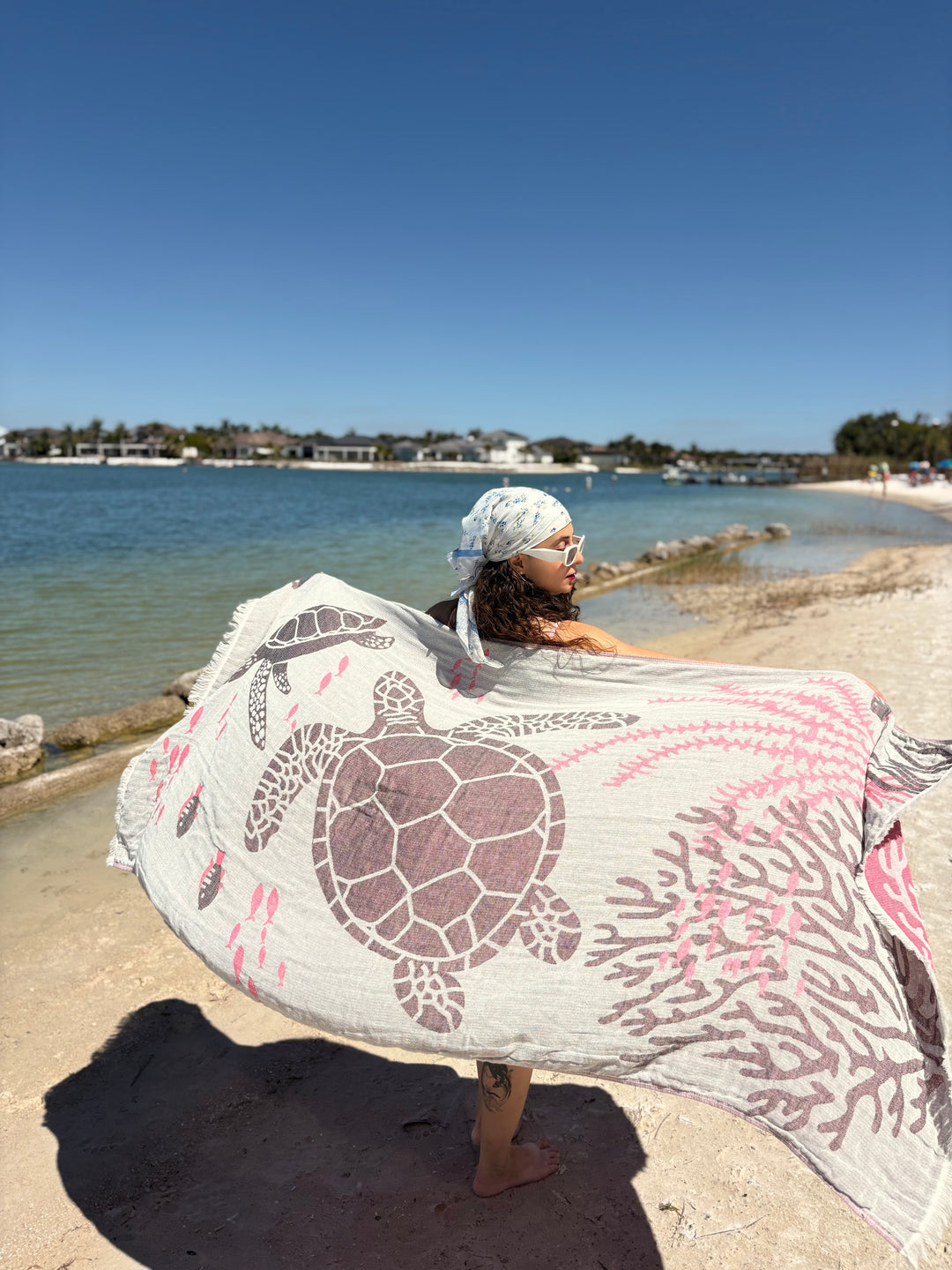 A woman stands on a sunny beach near the water, wearing a headscarf and holding the Turtle Turkish Towel by turkanhome.com, featuring jacquard-woven sea turtle and coral designs. Buildings and trees can be seen in the background.