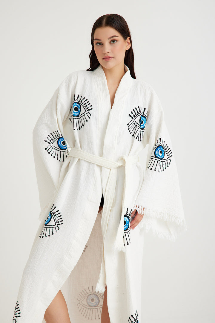 A woman poses against a white background in the Blue Eye With Lashes Kimono Robe by turkanhome.com, featuring a belt and intricate embroidered eye designs on the sleeves and front for a modern boho style.