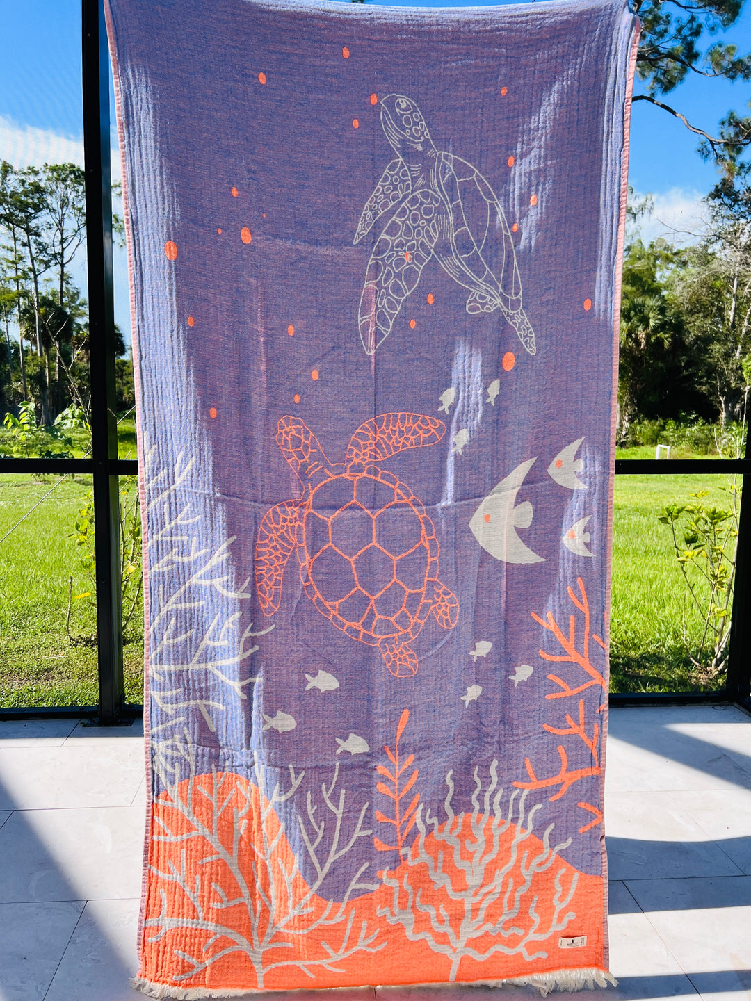 The Reef and Turtle Towel – Quick Drying Towel by turkanhome.com, a reversible blue and orange beach towel with sea turtle, fish, and coral designs, is displayed vertically indoors with a green lawn visible outside large windows.
