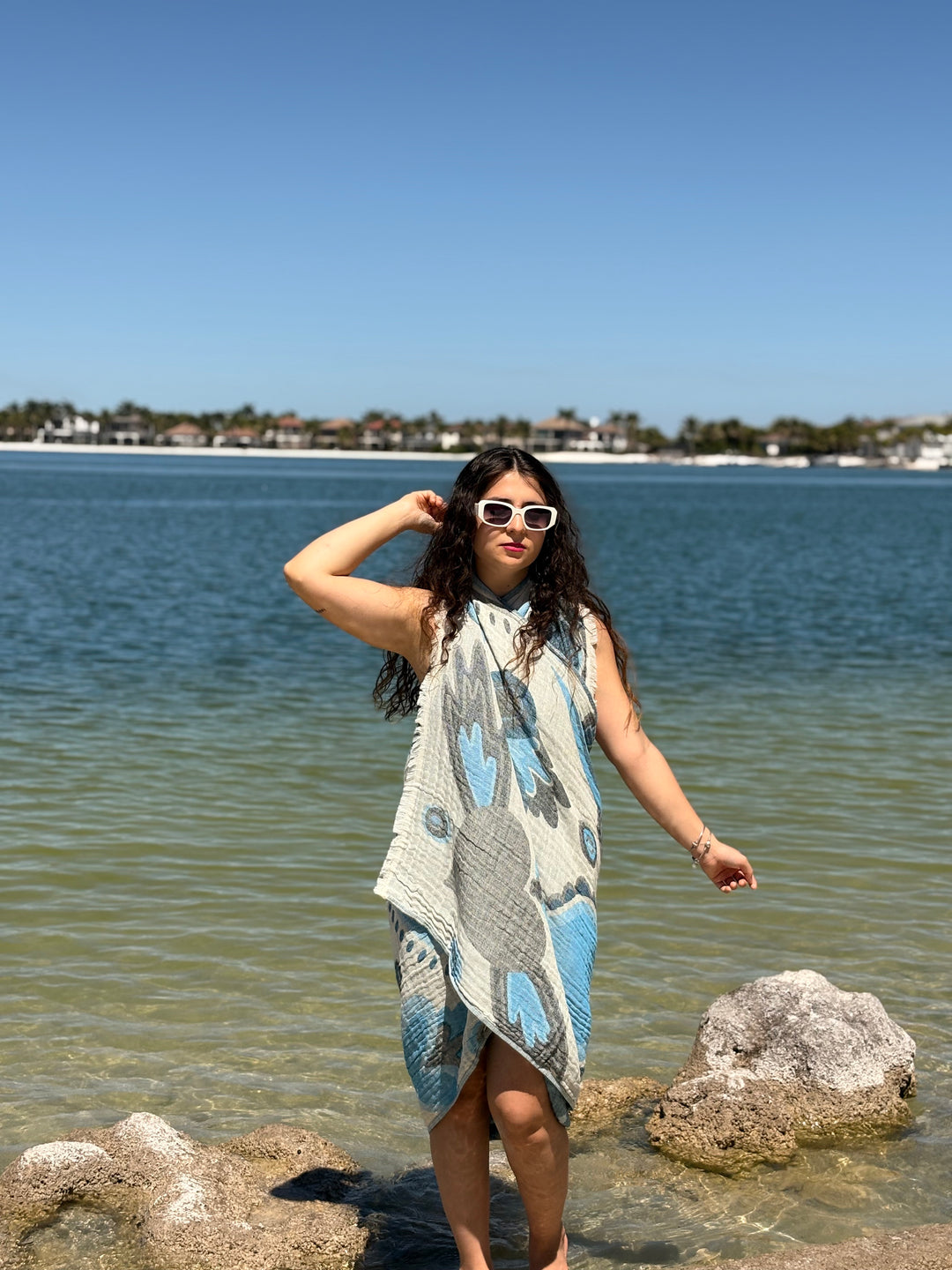 A woman in sunglasses and a patterned dress stands on lakeside rocks, draped in the turkanhome.com Heart Turkish Towel, with houses and trees across the water beneath a clear blue sky.