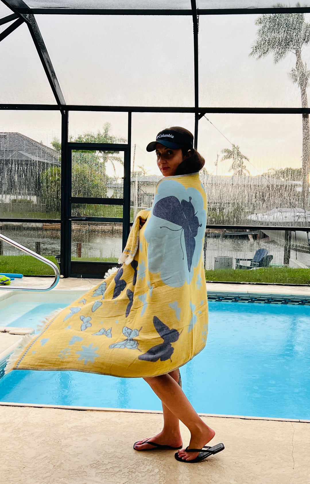 A woman by an indoor pool wears sunglasses and a black cap, wrapped in the yellow and blue Butterfly Towel – Quick Drying Towel by turkanhome.com. Palm trees and houses are seen outside through large windows.