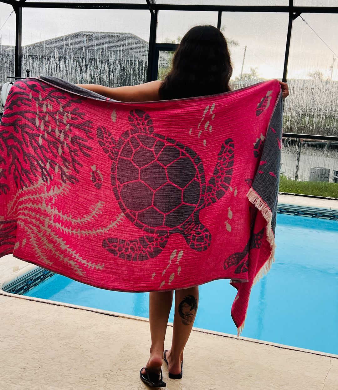 A person with long hair stands by a pool, holding up the turkanhome.com Reversible Sea Turtle Towel—a quick drying towel in bright pink featuring a large sea turtle. Facing away from the camera, they display a calf tattoo.