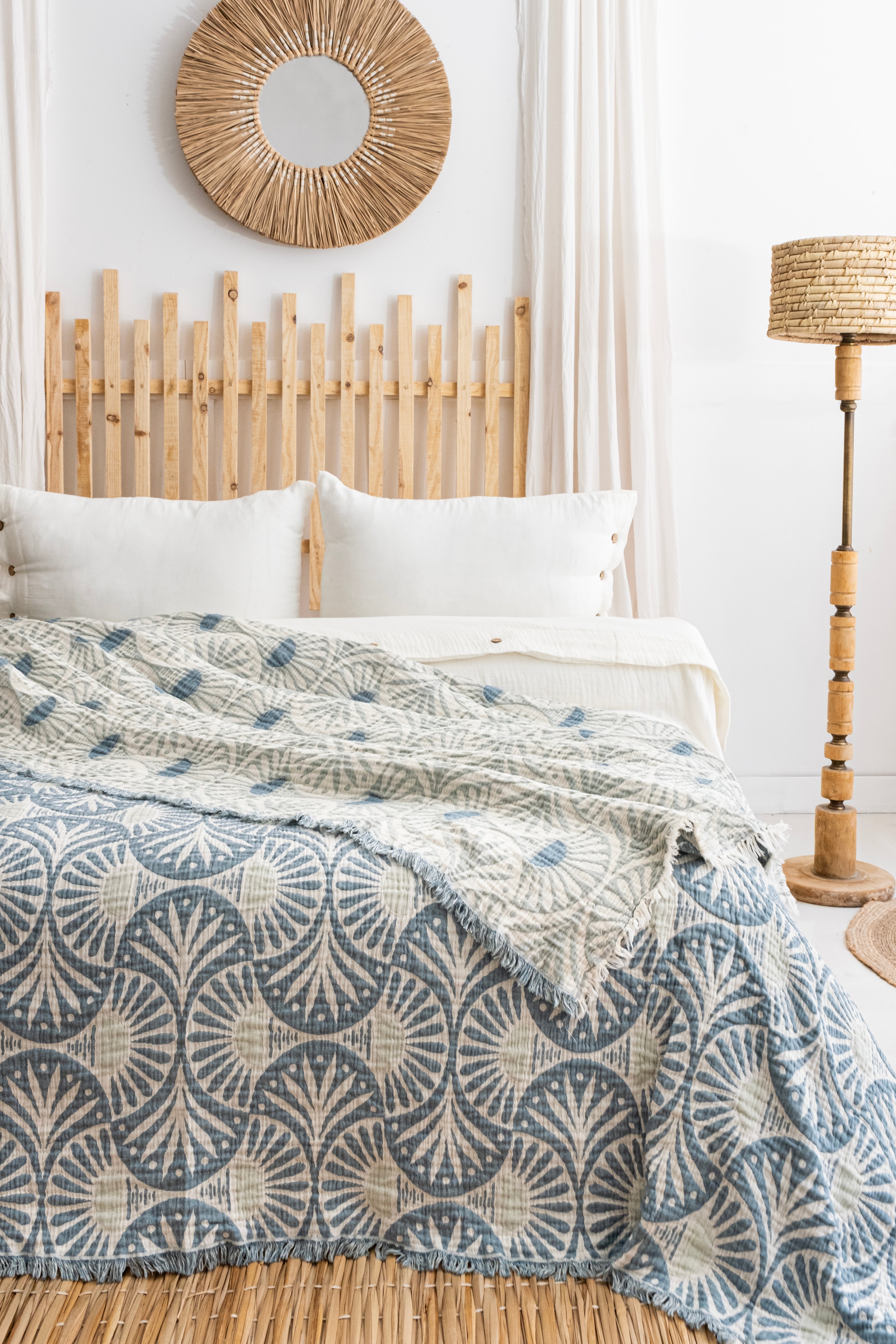 A cozy bedroom with a wooden headboard, white pillows, the Gingko Muslin Bed Blanket – Teal (King-Oversized Queen) from turkanhome.com, a round wicker mirror, and a wooden floor lamp with matching lampshade.