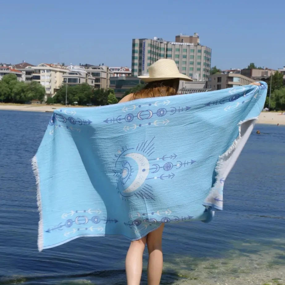 A person in a wide-brimmed hat stands by the water, wrapped in the Blue Seeing Eye Turkish Towel from turkanhome.com, featuring white and blue patterns. Buildings and trees are seen behind them under a clear sky.