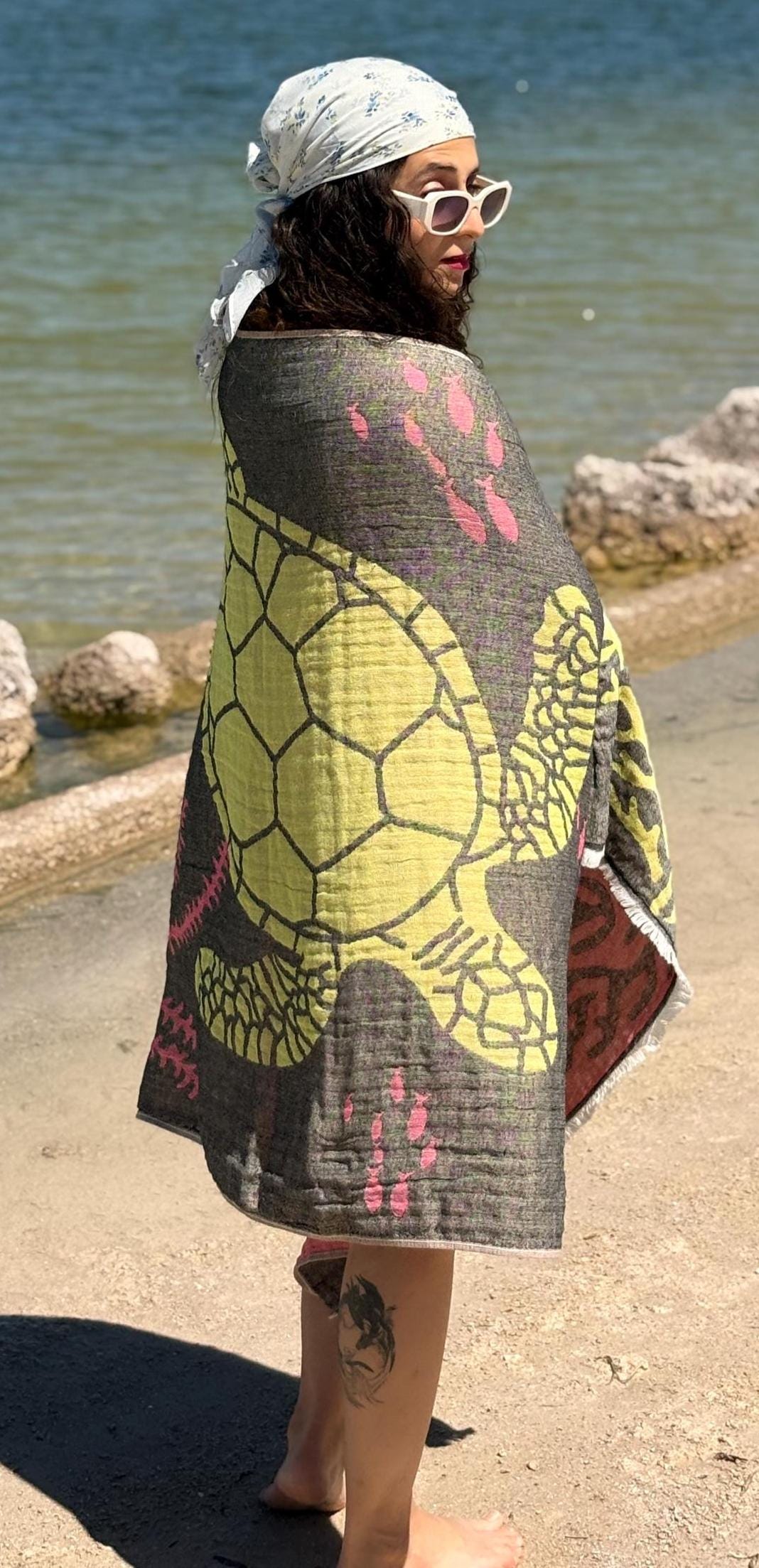 On a sandy beach, someone in white sunglasses and a bandana is wrapped in the turkanhome.com Reversible Sea Turtle Quick Drying Towel, with rocks and calm water behind them.
