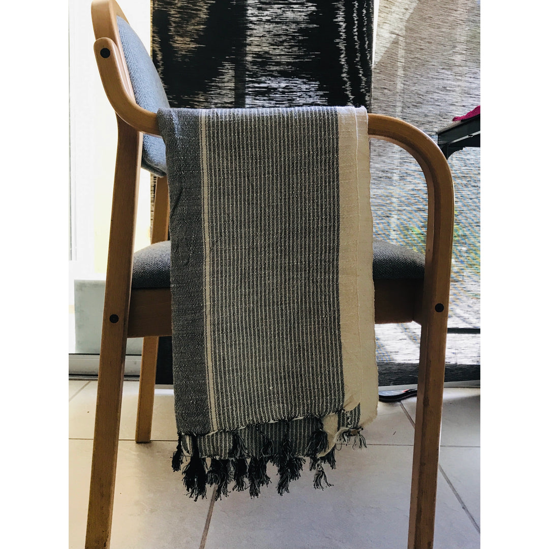 A wooden chair with a gray cushioned seat has a Grey Colorblock Peshtemal from turkanhome.com, featuring gray and white stripes, draped over its backrest, while a hand loomed towel hangs in the background.
