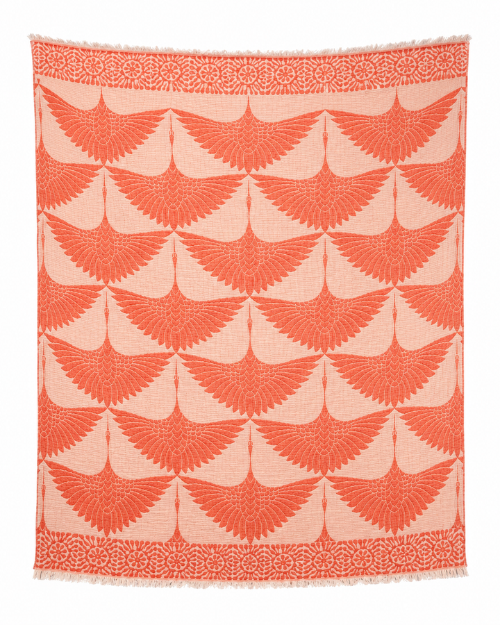 The Crane Muslin Throw Blanket Natural Border–Reversible Turkish Cotton from turkanhome.com is a rectangular textile featuring repeating orange winged motifs arranged in rows on a beige background, bordered by circular floral patterns along the top and bottom edges. Crafted from soft Turkish muslin cotton, this reversible throw is finished with fringe at the top and bottom for added texture and style.