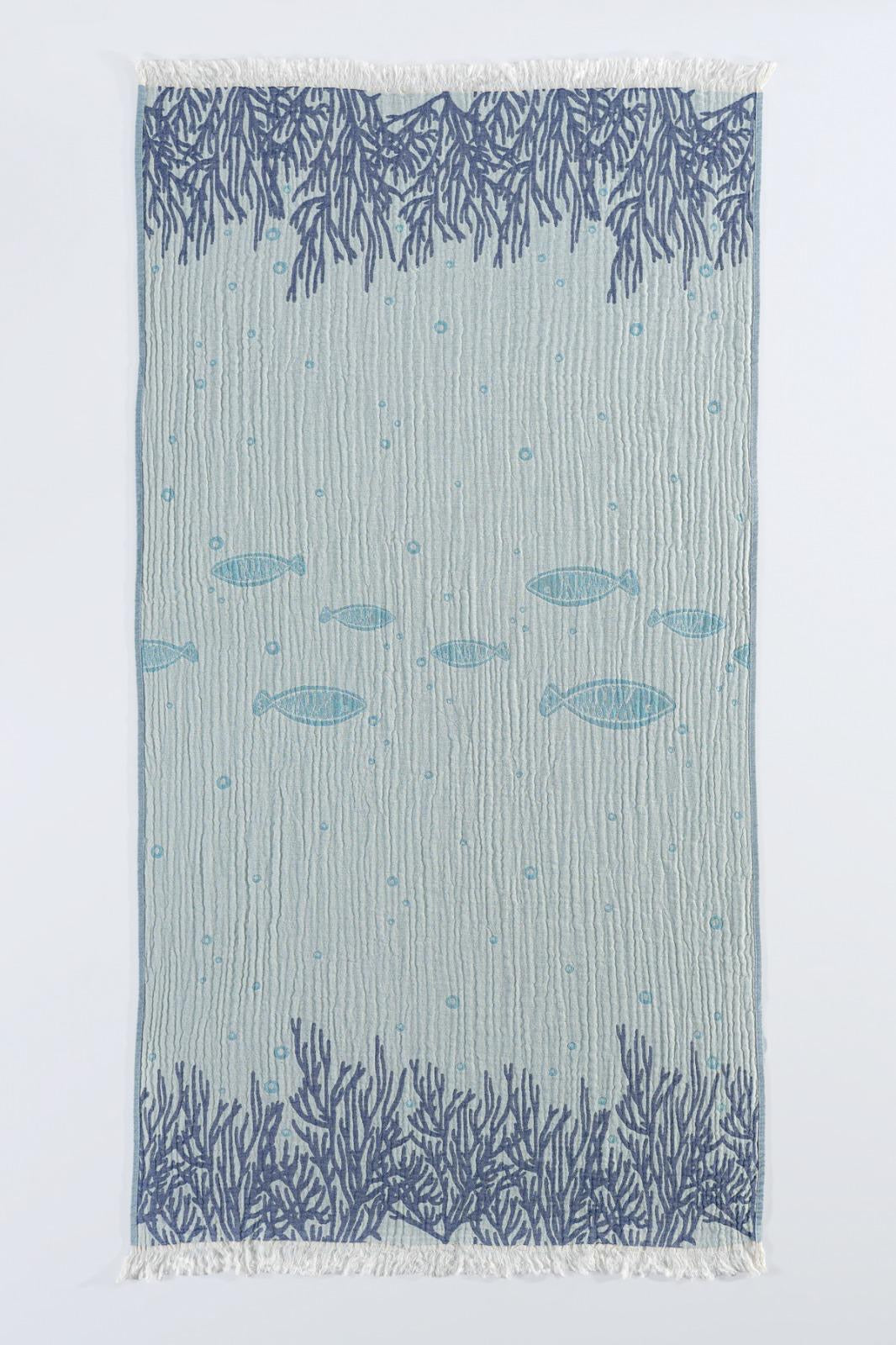 The Fishes and Coral Turkish Towel from turkanhome.com is jacquard woven from 100% cotton, featuring dark blue coral patterns on the edges, playful fish silhouettes in the center, and stylish fringed ends.