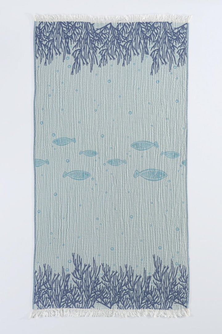 The Fishes and Coral Turkish Towel from turkanhome.com is jacquard woven from 100% cotton, featuring dark blue coral patterns on the edges, playful fish silhouettes in the center, and stylish fringed ends.