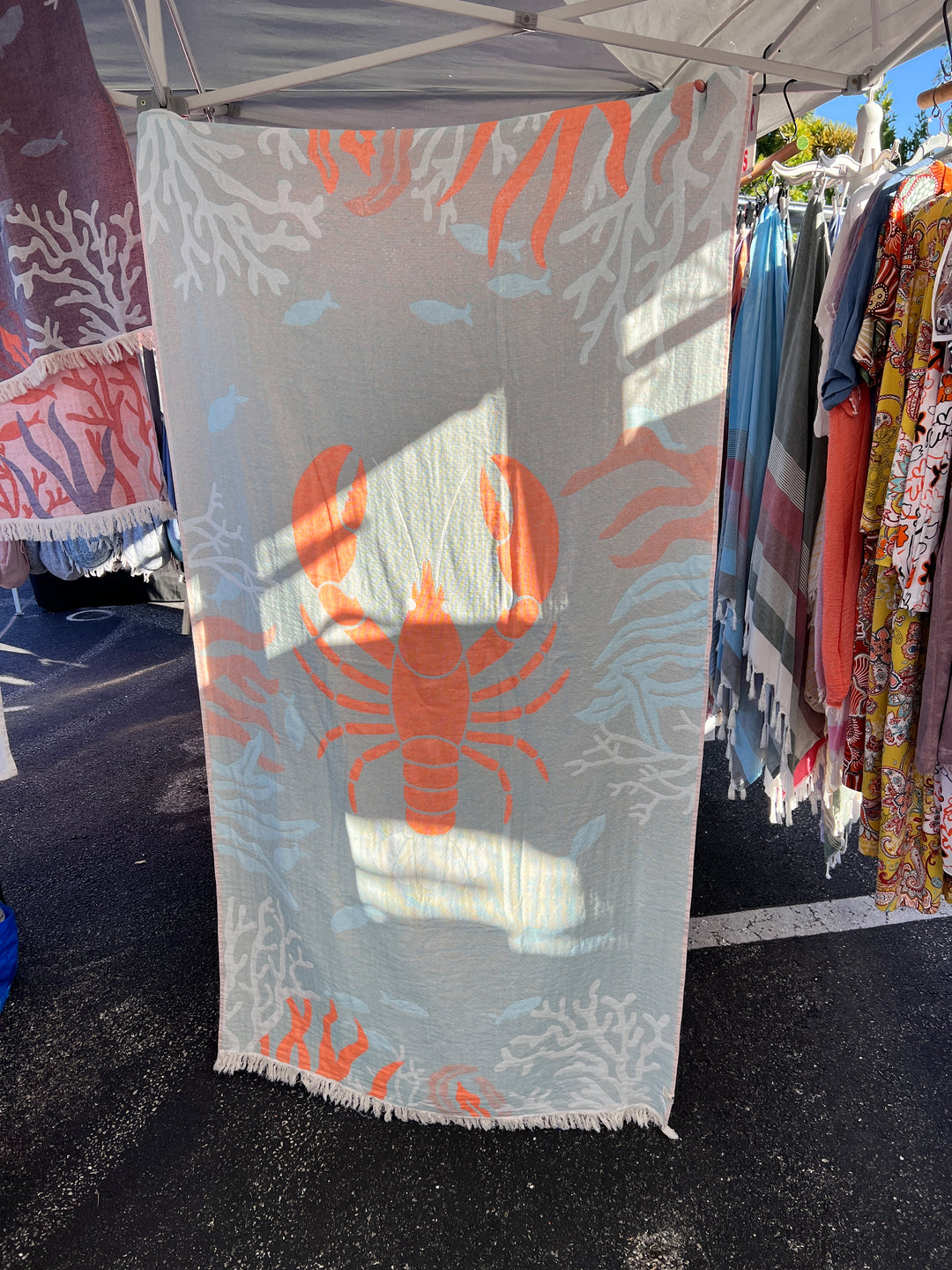 The Lobster Turkish Towel – Quick Drying Beach Towel from turkanhome.com, featuring an orange lobster and coral design, hangs at an outdoor market stall among other patterned fabrics and dresses in dappled sunlight.