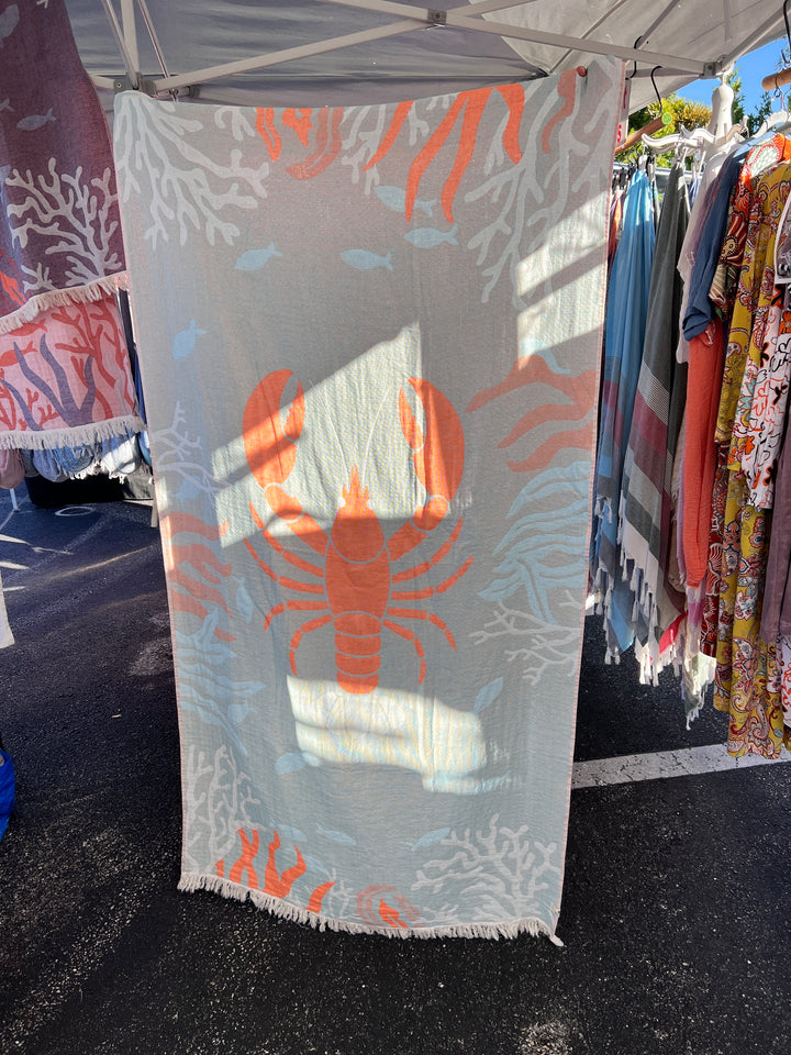 The Lobster Turkish Towel – Quick Drying Beach Towel from turkanhome.com, featuring an orange lobster and coral design, hangs at an outdoor market stall among other patterned fabrics and dresses in dappled sunlight.