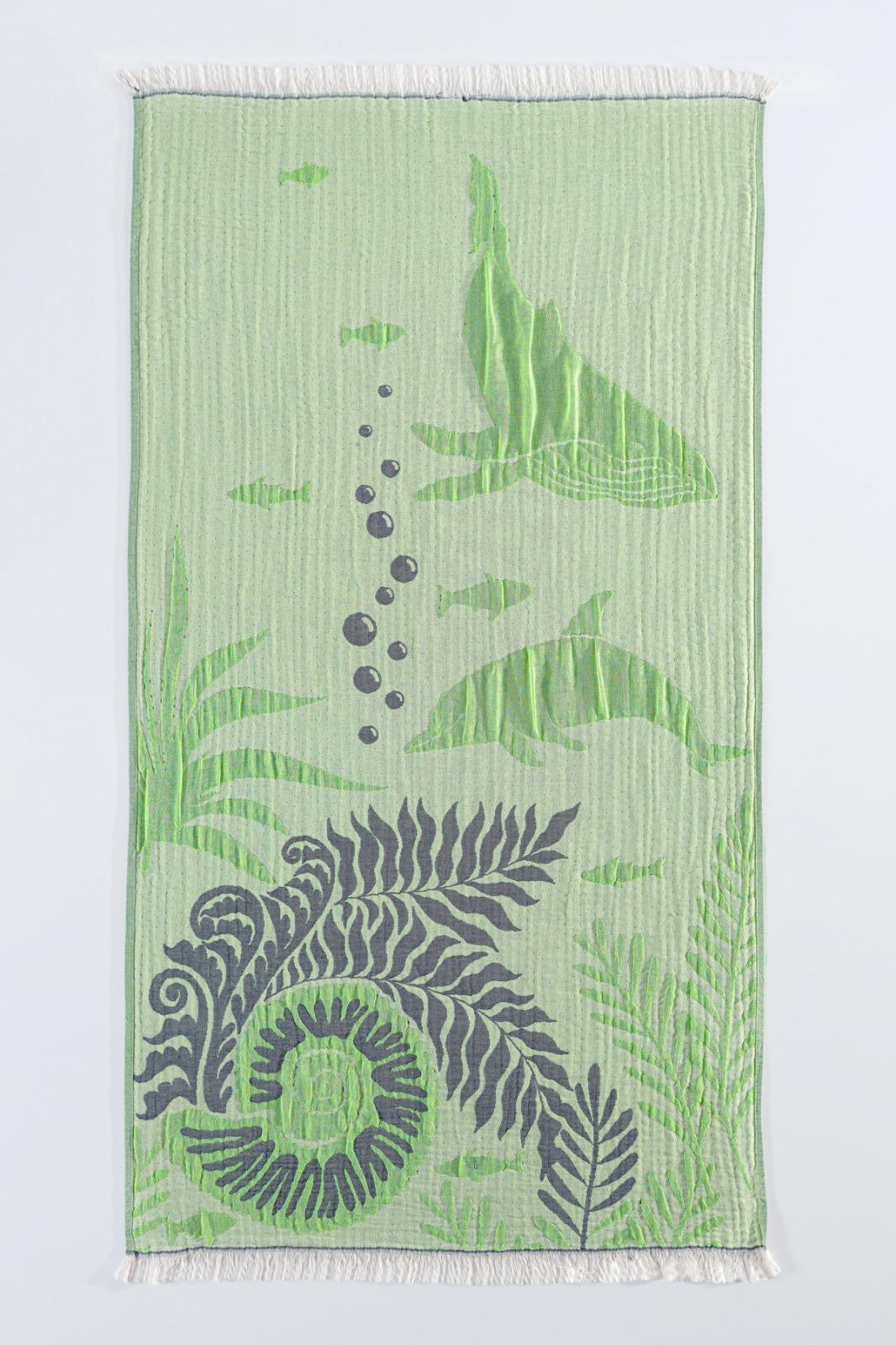 The Dolphin and Whale Turkish Towel by turkanhome.com is a light green 100% cotton towel featuring dolphins, small fish, bubbles, seaweed, a bold fern-like border, and white fringe on the short edges.