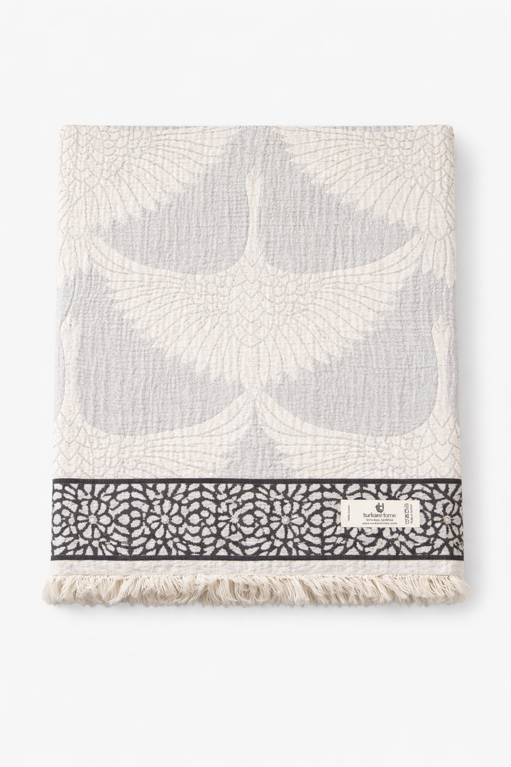 The Crane Muslin Throw Blanket – Reversible Turkish Cotton from turkanhome.com features a folded, textured gray and white design with crane motifs, a black-and-white decorative band near the edge, short fringe, and a branded label.