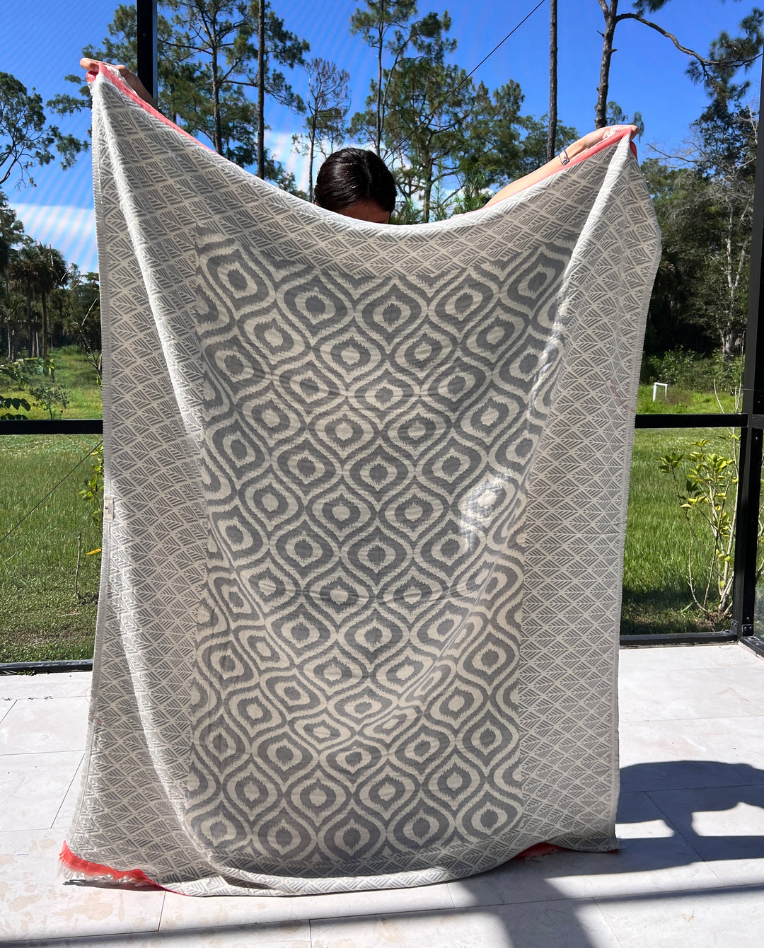 A person outdoors holds up the Ikat Throw from turkanhome.com, featuring a reversible geometric pattern in gray and white with a touch of red. Tall trees and greenery can be seen in the background through a mesh screen.