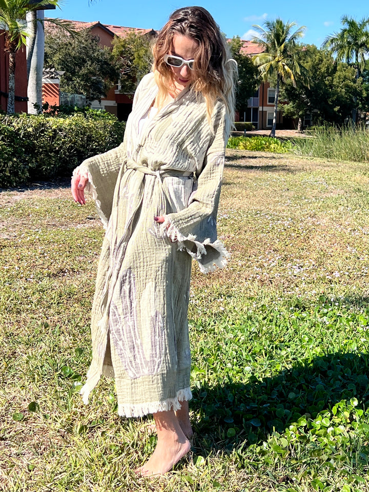 A woman in sunglasses and the Cactus Robe by turkanhome.com stands barefoot on grass in a sunny outdoor area with palm trees and buildings in the background—perfect as lounge or beach wear.