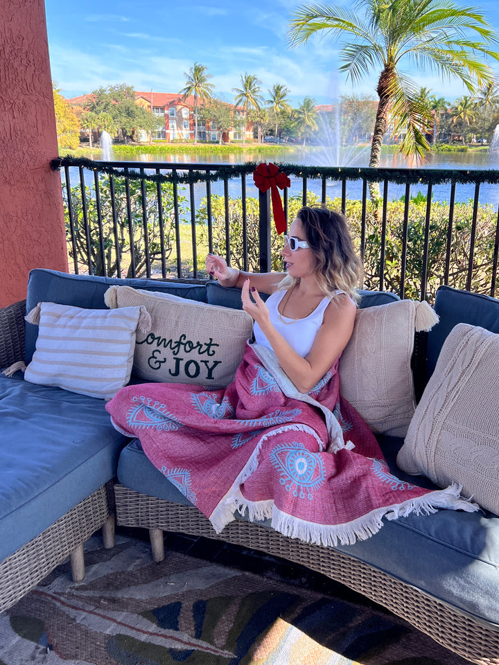 A woman in sunglasses relaxes on an outdoor sofa, wrapped in the Eye Red Sand Resistant Turkish Beach Towel from turkanhome.com, holding a cup. Palm trees, a lake, fountains, and a festive bow decorate the background. A pillow says Comfort & Joy.
