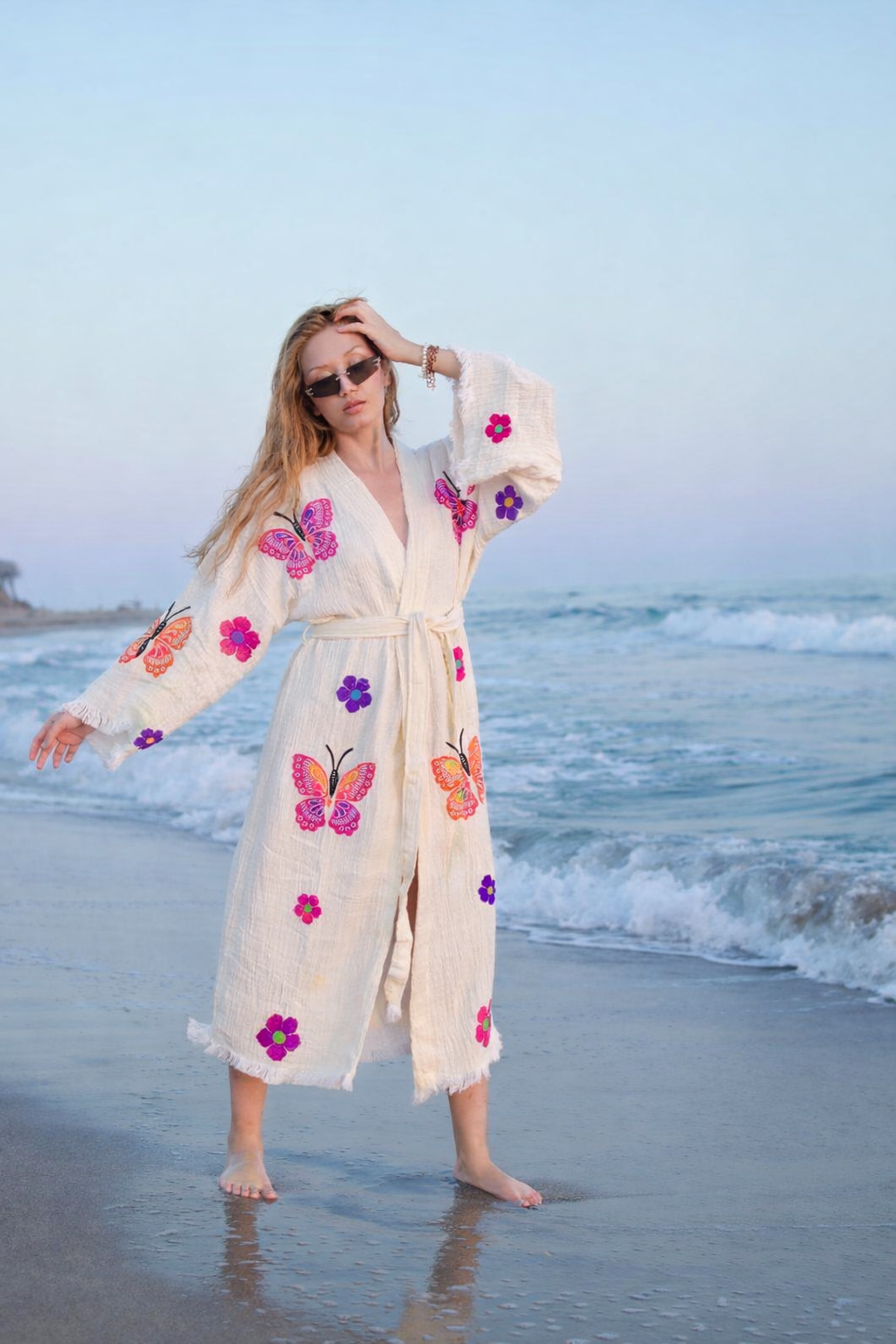 A woman in sunglasses and a "Give a Gift of Comfort" robe by turkanhome.com, featuring colorful butterfly and flower designs, stands barefoot on the beach with waves and a blue sky, capturing relaxed coastal style.