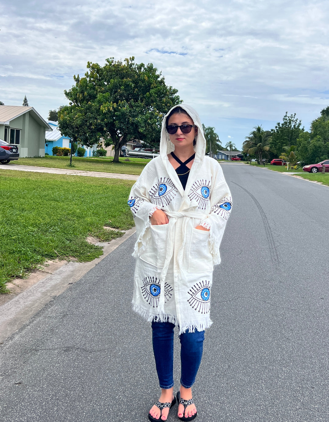 A person in sunglasses, a Black Top, jeans, sandals, and the Blue Eye With Lashes Kimono Robe by turkanhome.com stands on a suburban street with grass and trees under a cloudy sky.
