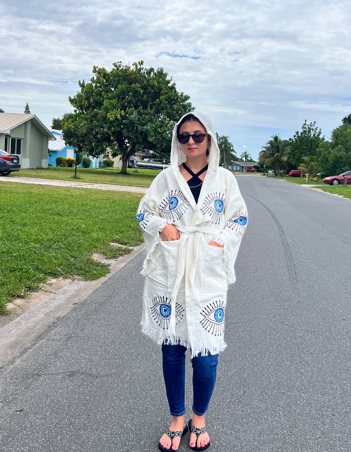 A person in sunglasses, a Black Top, jeans, sandals, and the Blue Eye With Lashes Kimono Robe by turkanhome.com stands on a suburban street with grass and trees under a cloudy sky.