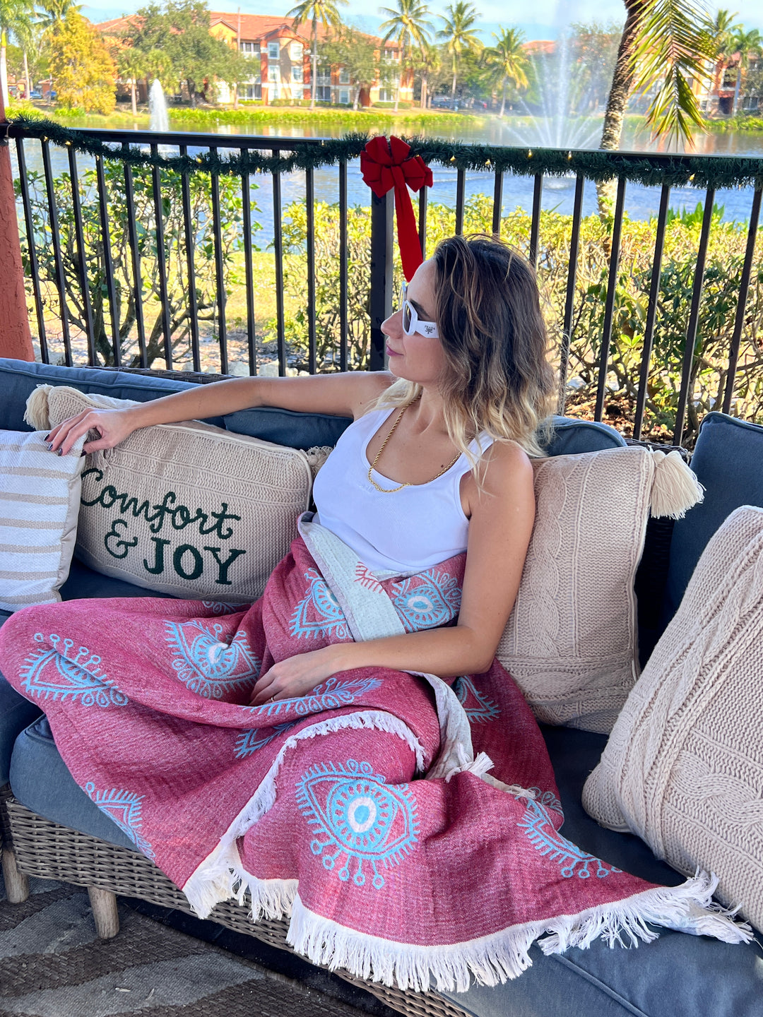 Relaxing on an outdoor couch by a lake, a woman in sunglasses and a white tank top is wrapped in the turkanhome.com Eye Red Sand Resistant Turkish Beach Towel, Throw, Shawl, surrounded by greenery and decorative pillows.