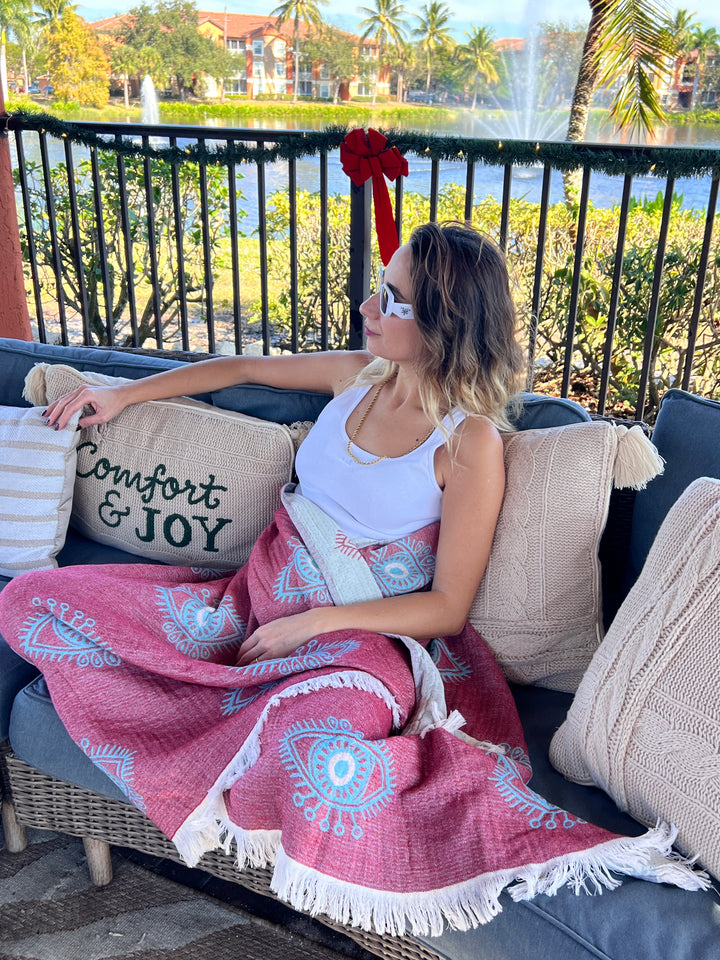 Relaxing on an outdoor couch by a lake, a woman in sunglasses and a white tank top is wrapped in the turkanhome.com Eye Red Sand Resistant Turkish Beach Towel, Throw, Shawl, surrounded by greenery and decorative pillows.