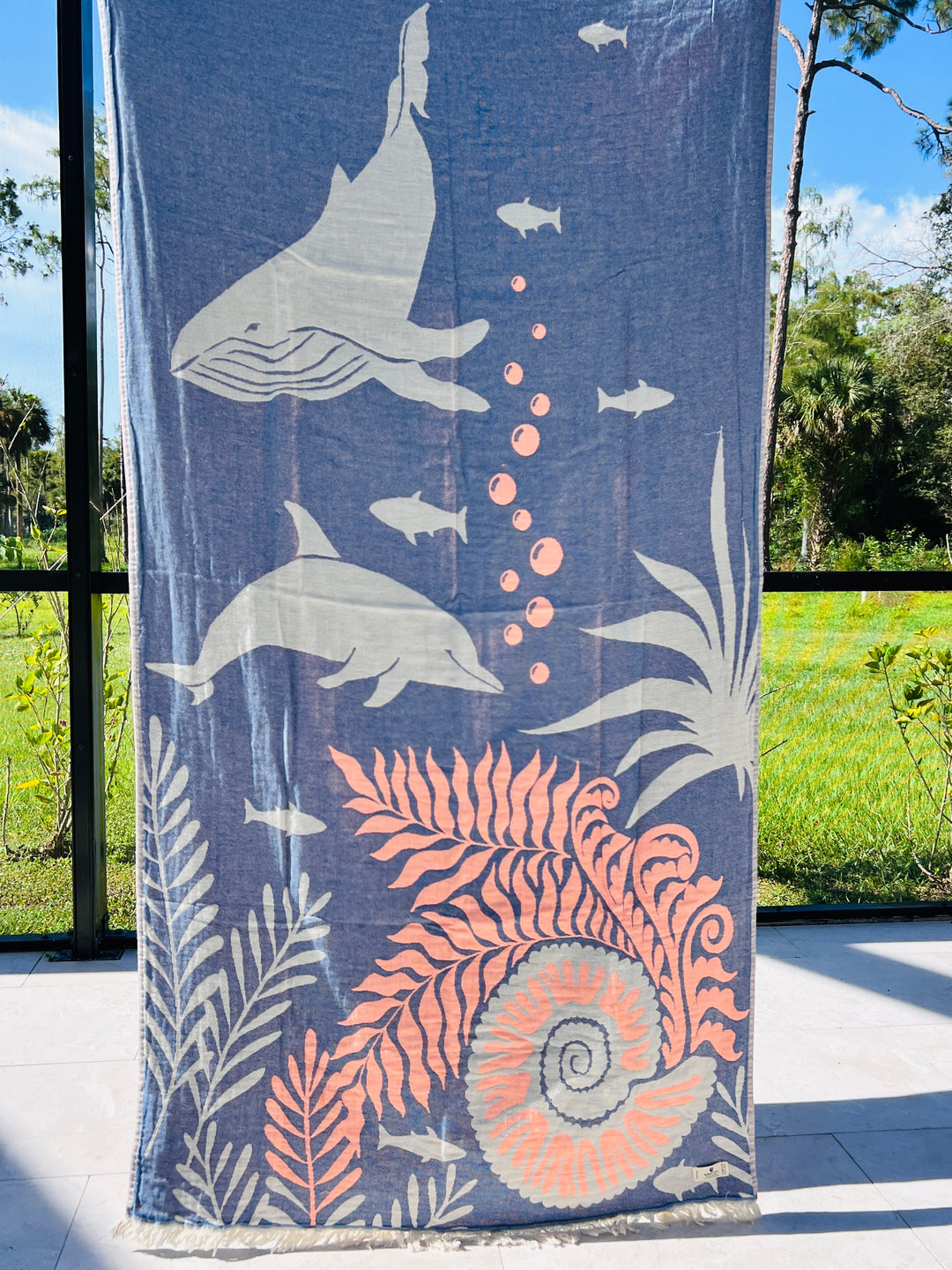 The Dolphin and Whale Turkish Towel by turkanhome.com hangs vertically, jacquard woven from 100% cotton with light whale, dolphin, fish, plant, bubble, and seashell illustrations. Sunlight and greenery glow through the window behind it.
