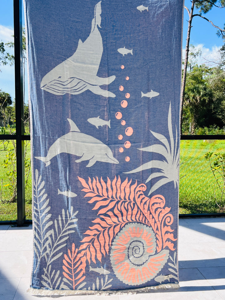 The Dolphin and Whale Turkish Towel by turkanhome.com hangs vertically, jacquard woven from 100% cotton with light whale, dolphin, fish, plant, bubble, and seashell illustrations. Sunlight and greenery glow through the window behind it.