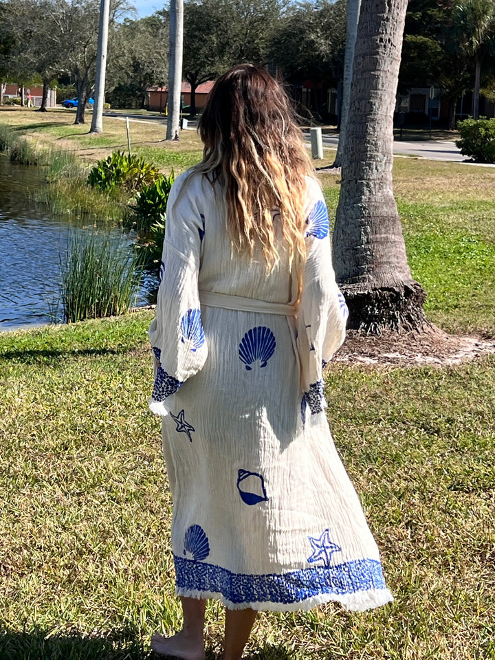 A woman with wavy hair stands by a lake wearing the Sea Shell Kimono Robe-Blue from turkanhome.com, which features blue seashell, starfish, and wave designs. She faces away, with trees and houses in the background.
