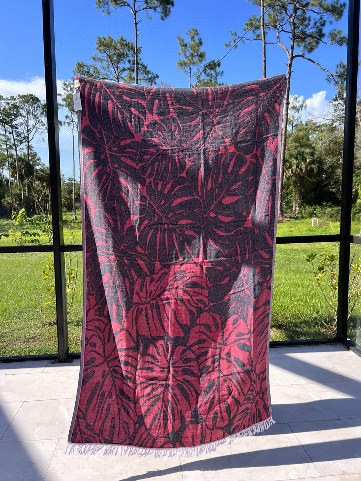 The Botanical Turkish Towel by turkanhome.com, featuring a pink and gray monstera leaf design, hangs in front of a screened porch with green grass, trees, and blue sky in the background.