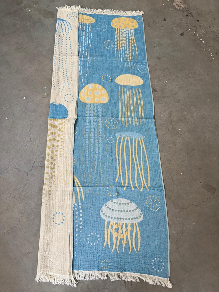 The Jellyfish Reversible Turkish Beach Towel by turkanhome.com features blue, yellow, and white jellyfish on a blue background. Quick drying and ideal for the beach, it showcases its lighter reverse side with a folded edge on a concrete floor.