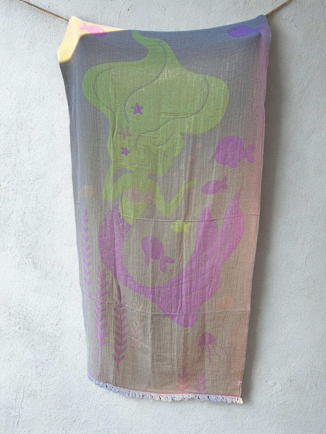 The Mermaid Turkish Towel by turkanhome.com, made from 100% cotton and featuring a pastel mermaid with fish and sea plants, hangs on a string against a light gray textured wall.