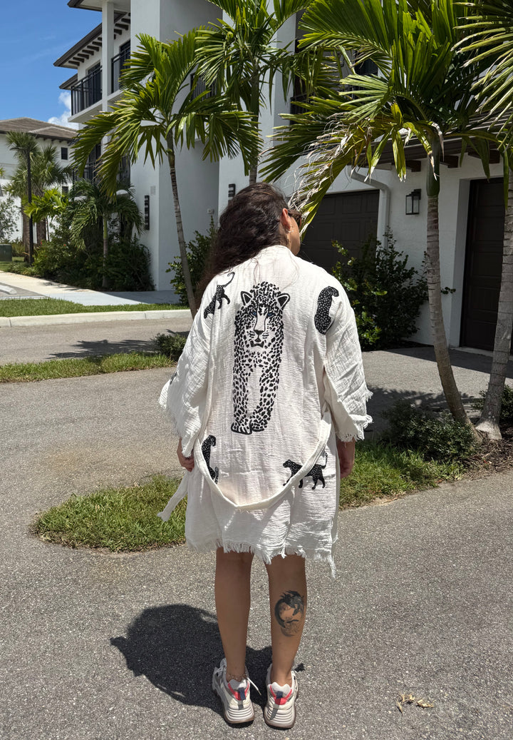 A person with long hair stands outdoors, facing away, wearing a light-toned Leopard Cotton Kimono Robe-Short from turkanhome.com, featuring a bold cheetah graphic and black cat accents. Palm trees and modern houses can be seen in the background, and the person has a tattoo on their right calf.