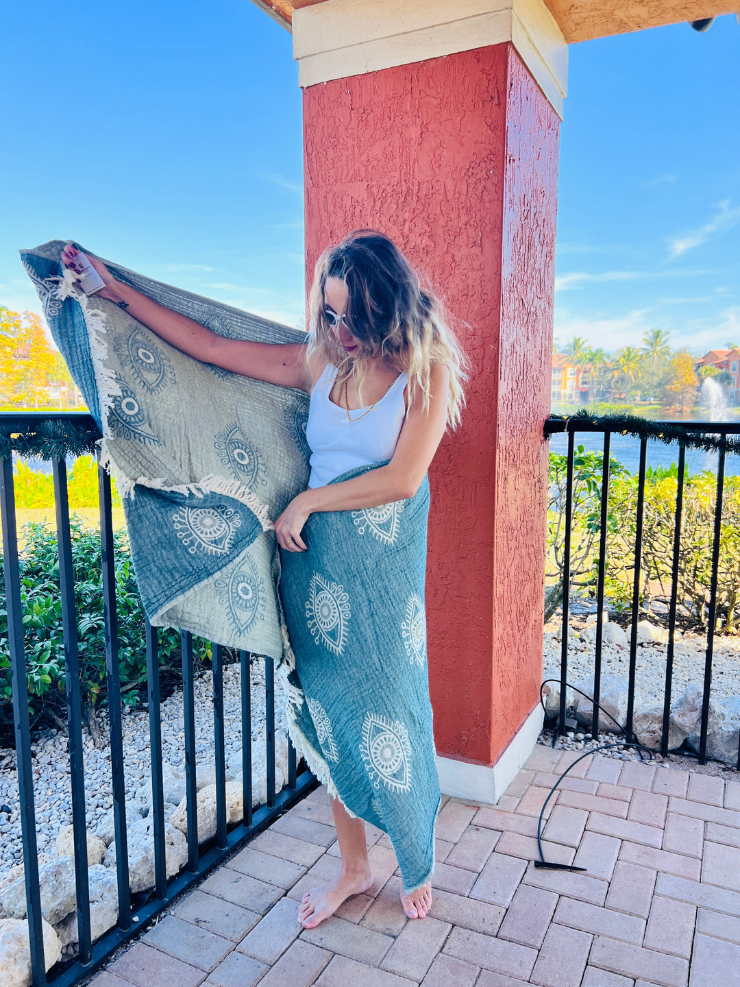 A woman in sunglasses and a white tank top stands barefoot on a patio, draping the Eye Sand Resistant Turkish Beach Towel by turkanhome.com—made from recycled fibers—around herself. A red column, black railing, greenery, and water are visible behind her.