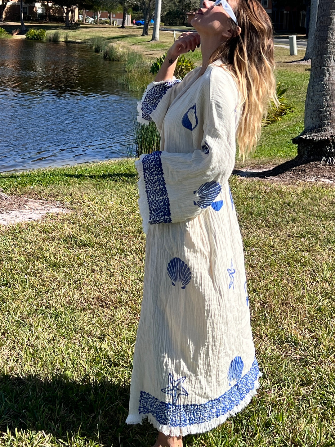 A woman stands barefoot on grass by a lake, wearing the Sea Shell Kimono Robe-Blue from turkanhome.com—its long, cream-colored muslin cotton fabric features blue seashell and starfish patterns. Sunlight illuminates her as she looks up, touching her chin.
