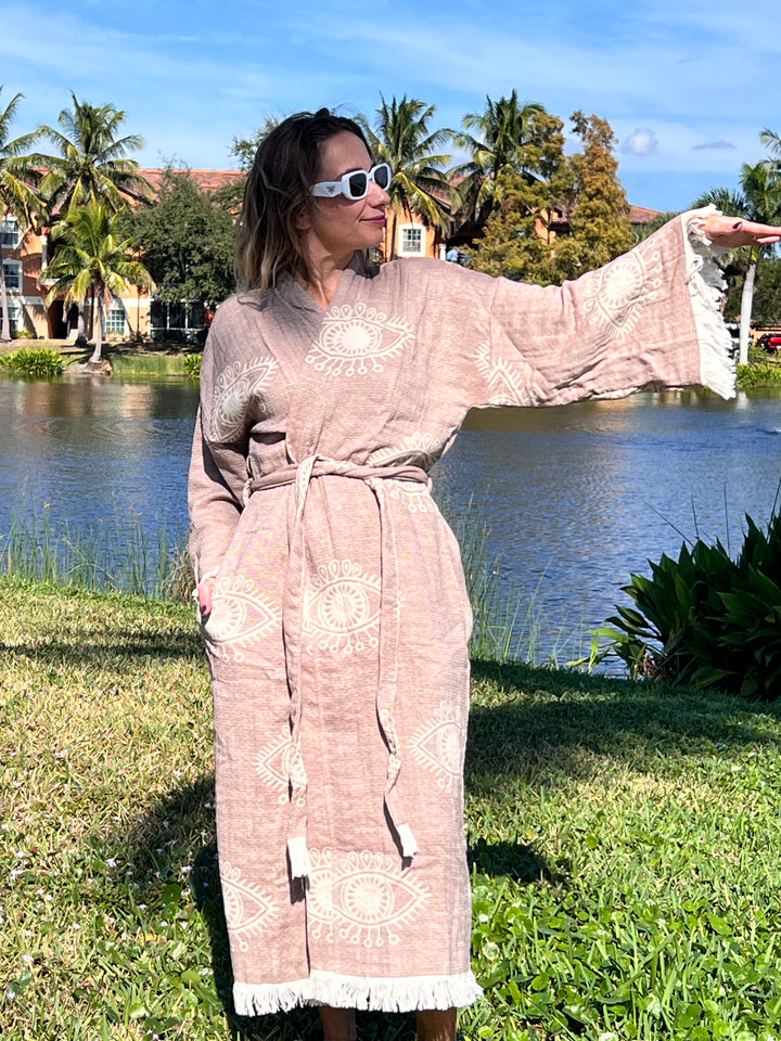 A woman wearing the Brown Eye Robe Kimono from turkanhome.com stands on grass near a lake, one arm extended. She wears sunglasses; palm trees and buildings appear in the background under a blue sky.
