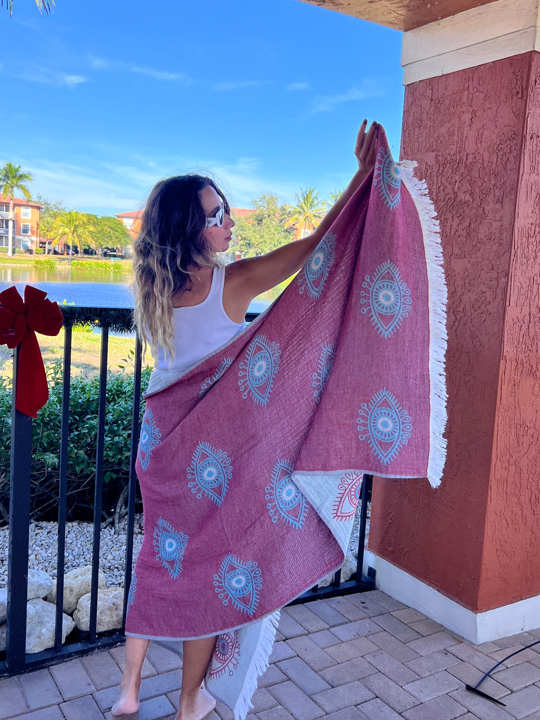 A woman in sunglasses and a white tank top displays the Eye Sand Resistant Turkish Beach Towel by turkanhome.com, featuring red, white, and blue patterns, while standing on a patio with palm trees and a pond in the background.