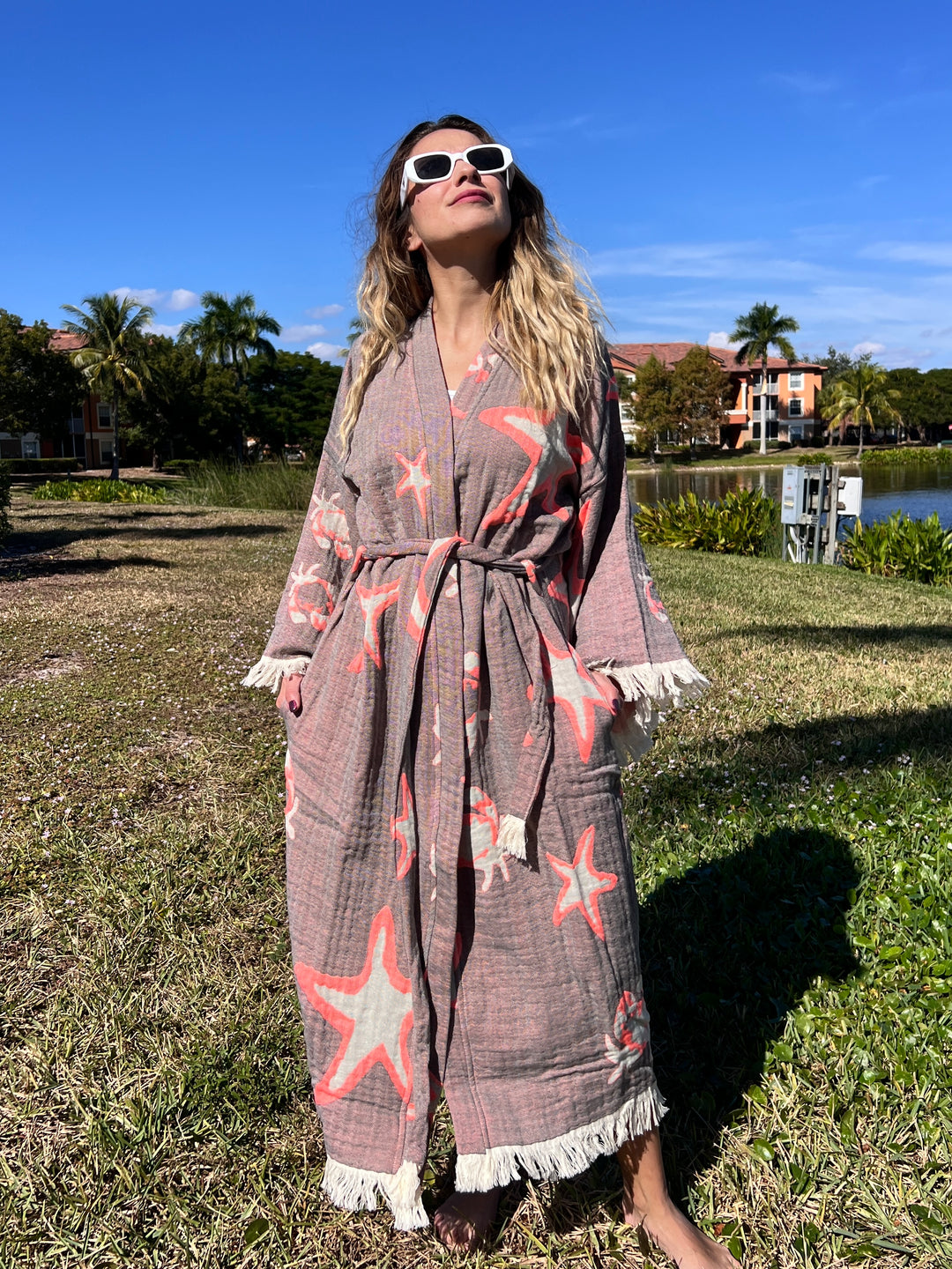A woman in the Pink Sea Star Robe by turkanhome.com, a boho kimono with starfish patterns and white fringe, stands on grass by a lake, smiling in sunglasses beneath a clear blue sky with palm trees and buildings behind her.