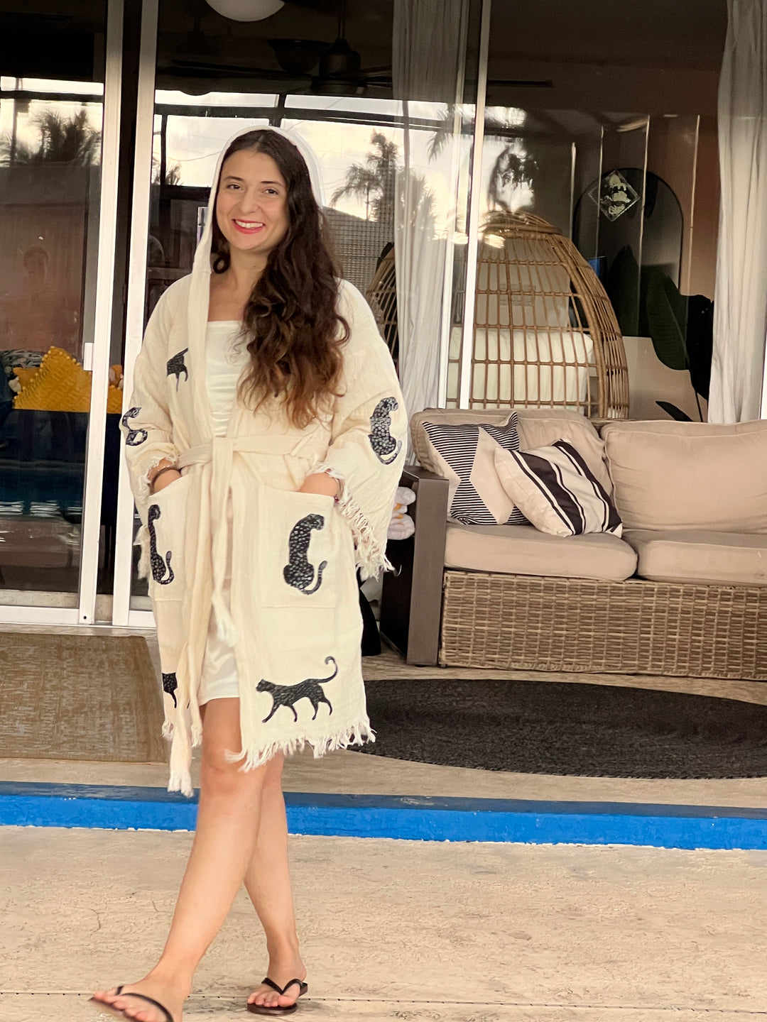 A woman with long brown hair in a white dress and a Black Leopard Hooded Cotton Kimono Robe by turkanhome.com stands barefoot on a patio, smiling near wicker furniture and sliding glass doors.
