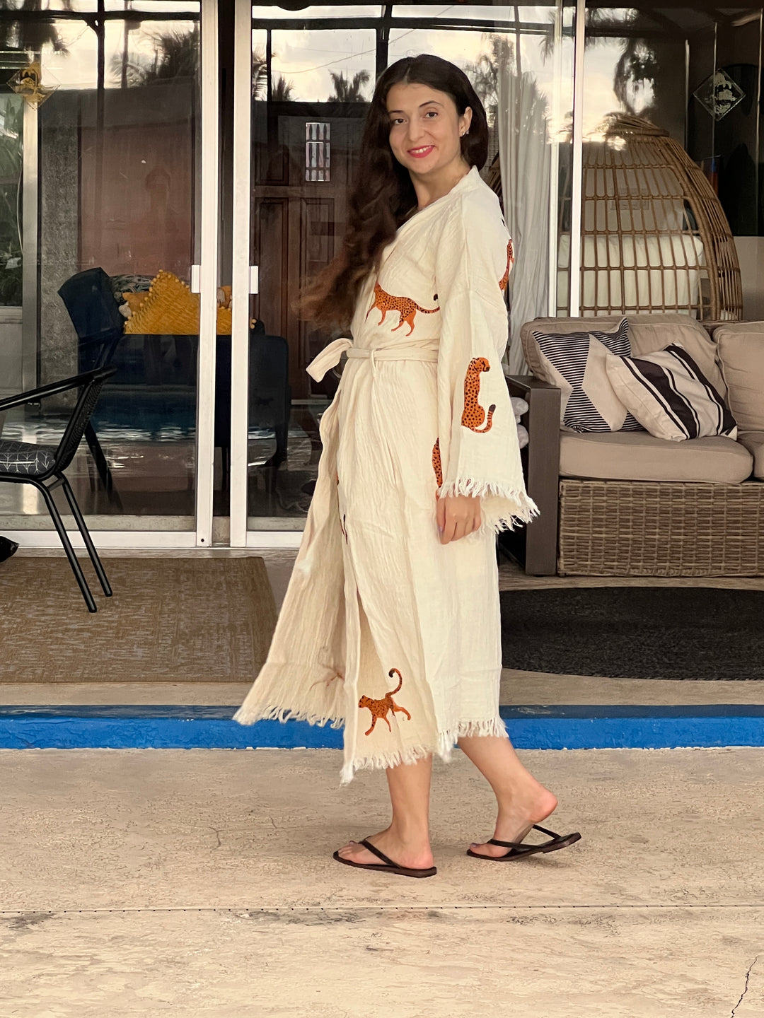 A woman with long brown hair, wearing the Orange Leopard Cotton Kimono Robe by turkanhome.com and black sandals, stands outside on a patio furnished with wicker and a hanging chair.