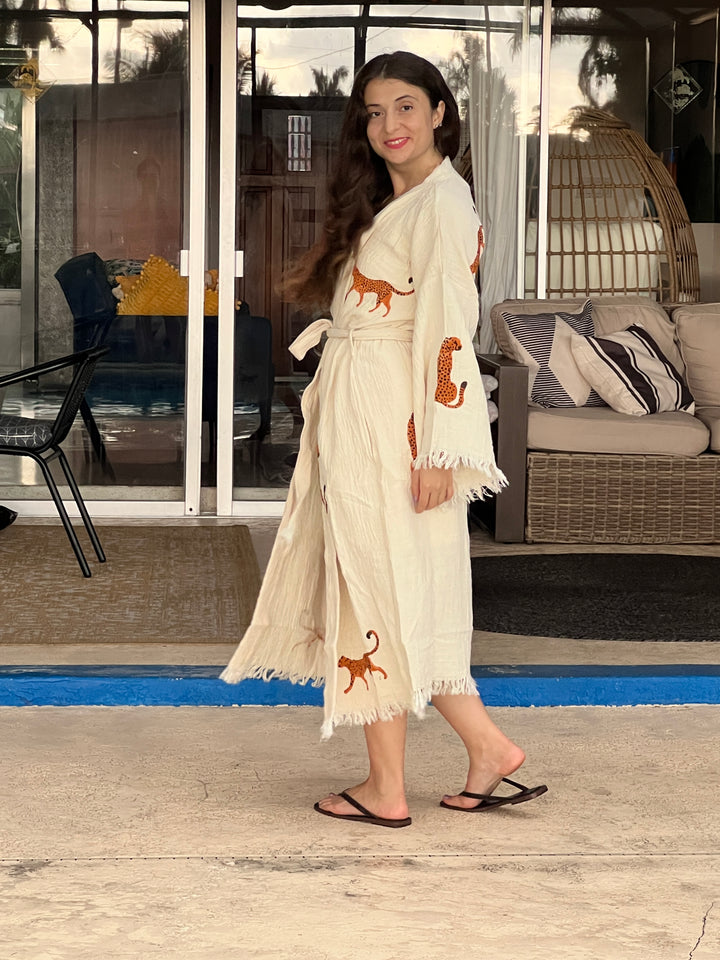A woman with long brown hair, wearing the Orange Leopard Cotton Kimono Robe by turkanhome.com and black sandals, stands outside on a patio furnished with wicker and a hanging chair.