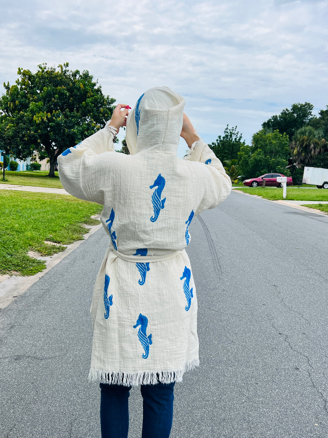 A person stands on a suburban street, facing away and adjusting the hood of a turkanhome.com Seahorse Kimono Robe with Hoodie, featuring eco-friendly fabric and blue seahorse prints, with trees and houses in the background.
