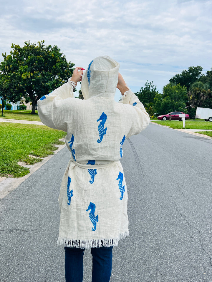 A person stands on a suburban street, facing away and adjusting the hood of a turkanhome.com Seahorse Kimono Robe with Hoodie, featuring eco-friendly fabric and blue seahorse prints, with trees and houses in the background.