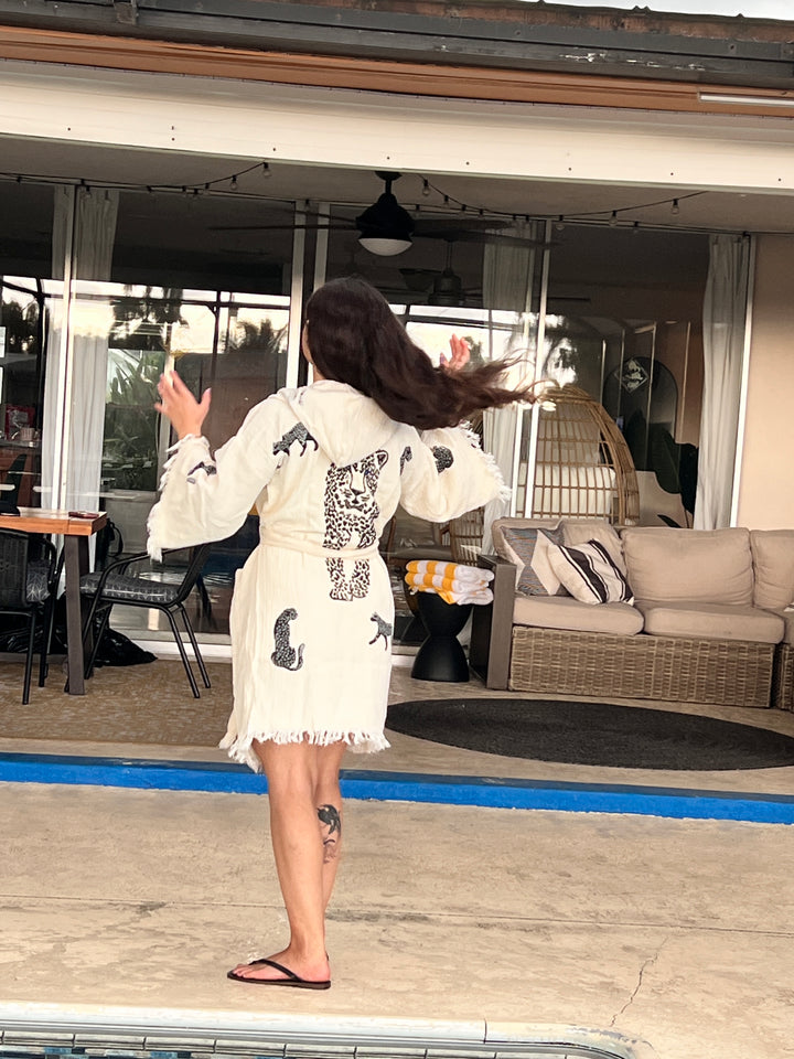 A person with long dark hair, wearing a Black Leopard Hooded Cotton Kimono Robe by turkanhome.com, stands by a pool facing away. Their hair swings, showing a left leg tattoo. Patio furniture is visible in the background.