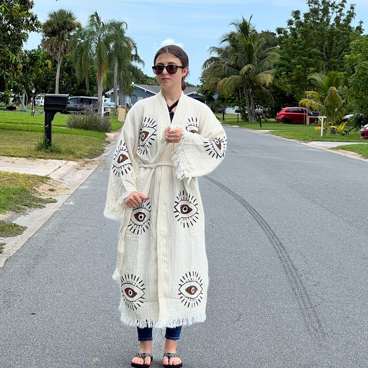 A woman wearing sunglasses and a Brown Eye Kimono Robe by turkanhome.com stands on a palm-lined suburban street with parked cars.