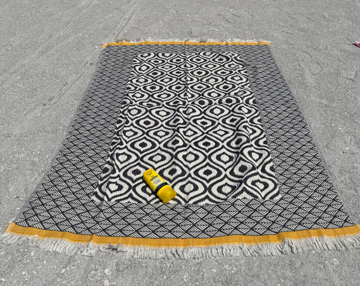 An Ikat Throw from turkanhome.com with a reversible geometric pattern and yellow trim lies on the gray sand. A yellow bottle of Banana Boat sunscreen sits on the throw.