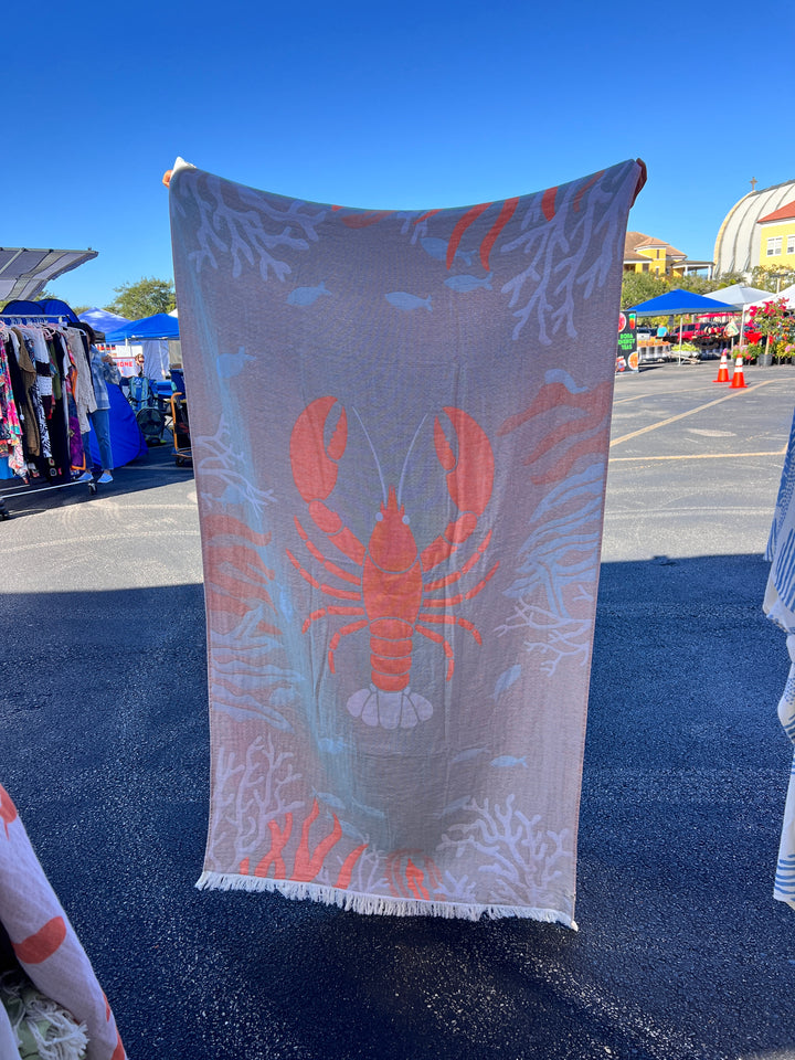 The Lobster Turkish Towel – Quick Drying Beach Towel by turkanhome.com, featuring a vibrant orange lobster with coral and fish designs, is displayed at an outdoor market beneath a clear blue sky, with booths and tents in the background.