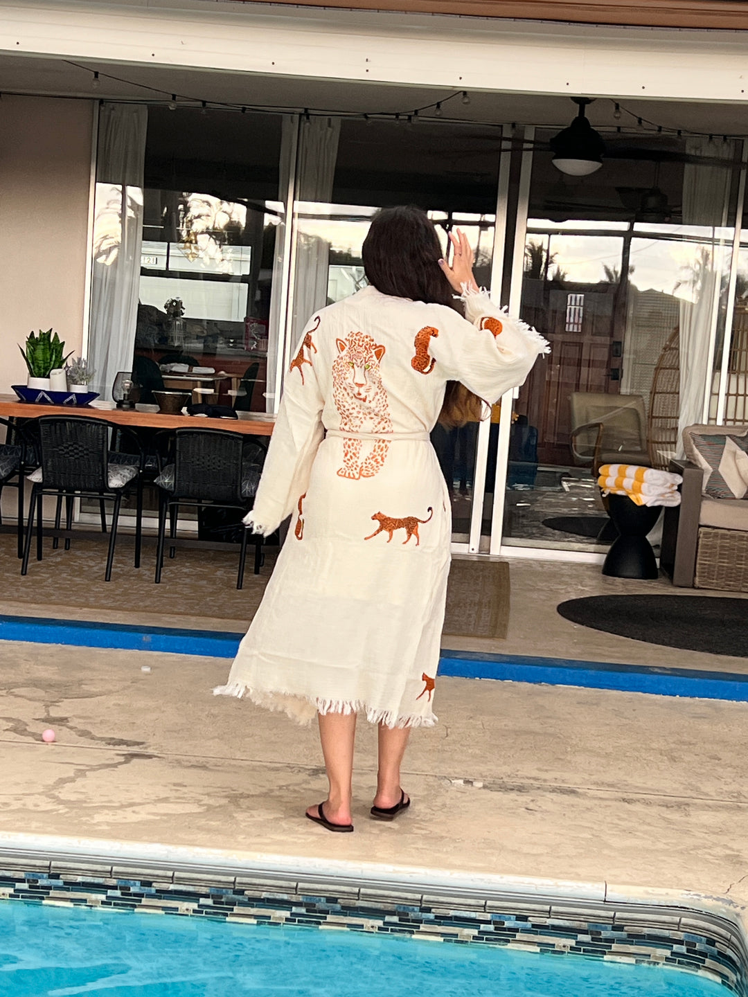 A woman with long brown hair stands by a pool, facing away, wearing the Orange Leopard Cotton Kimono Robe beach cover up from turkanhome.com. She raises her right hand; patio furniture and a glass door are visible in the background.