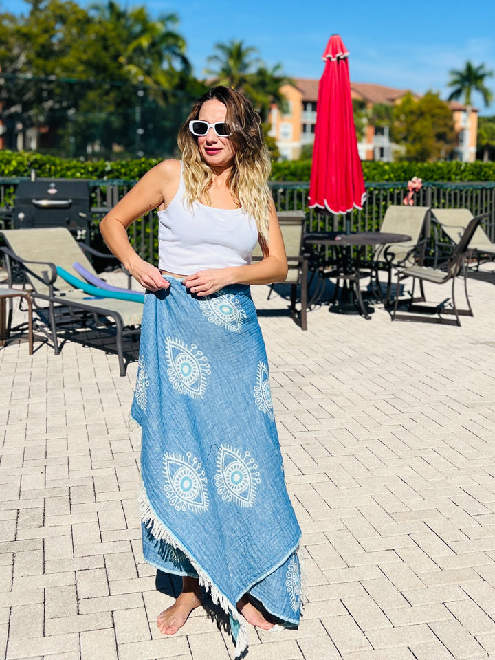 A woman stands barefoot poolside, wearing sunglasses, a white tank top, and the Eye Blue Sand Resistant Turkish Beach Towel by turkanhome.com as a sarong. Lounge chairs, tables, a red umbrella, palm trees, and sunny skies complete the scene.