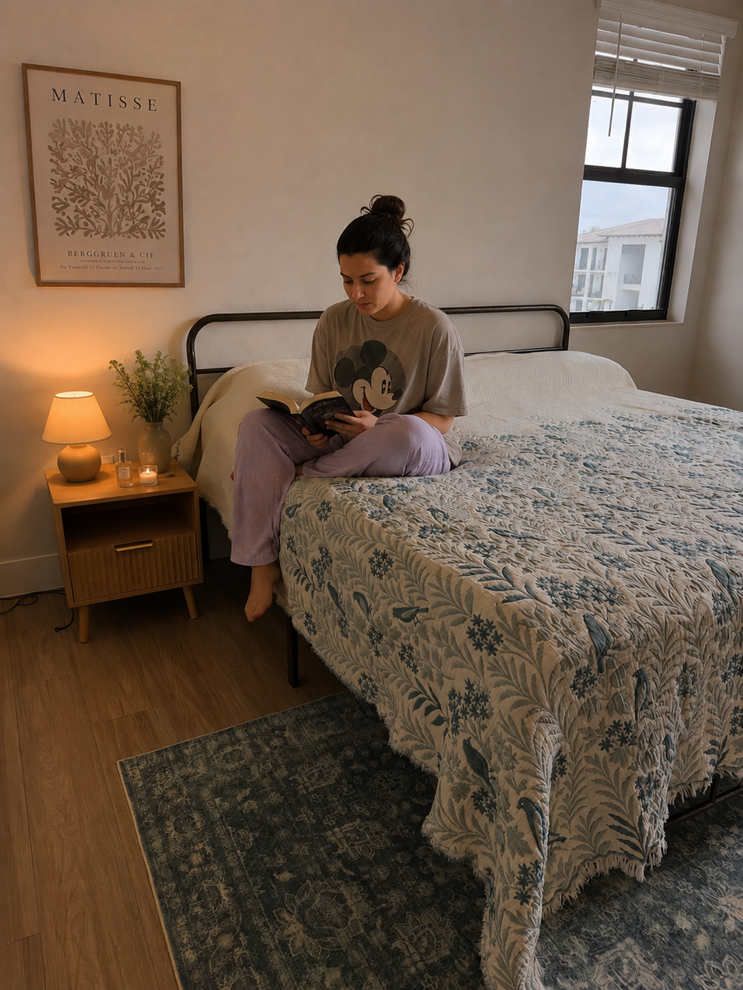 A woman in pajamas sits on a bed reading a book in a cozy, softly lit bedroom adorned with the turkanhome.com Luxury 7-Layer Cotton Muslin Quilted Blanket in Mint Bird, complemented by a nightstand, lamp, Matisse poster, and a window revealing a building outside.